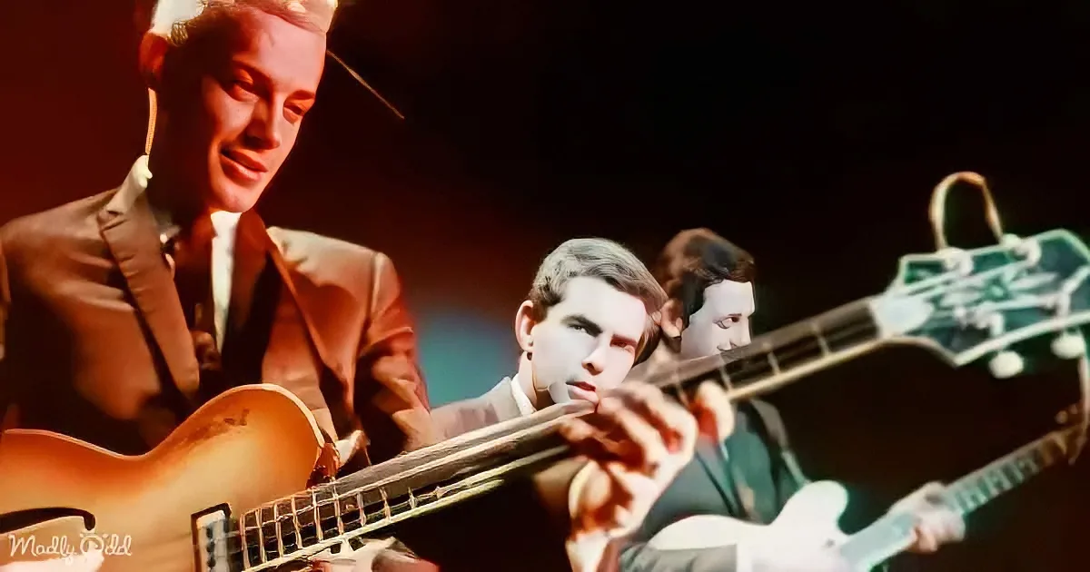 A man in a suit plays a guitar on stage, with another musician in the background.