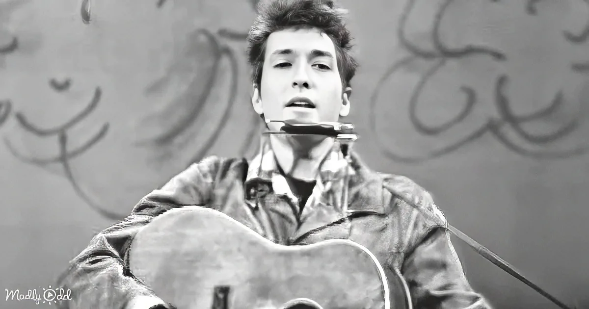 A person plays guitar and harmonica, wearing a jacket, against a wall with graffiti.