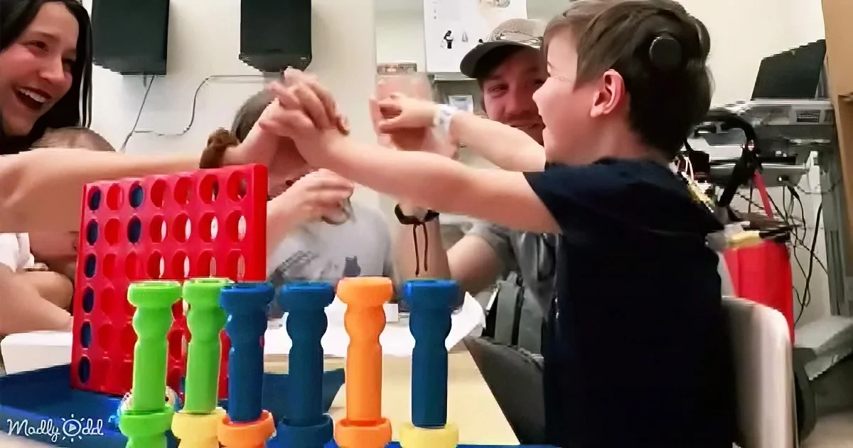 A boy in a black shirt is enthusiastically playing a game with colorful pegs and a red board, surrounded by smiling adults.