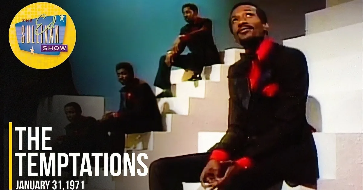 A man in a black suit sits on a white chair, singing passionately, with four other men in similar attire sitting on steps...