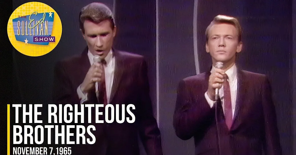 Two men in suits sing into microphones, one with eyes closed, against a dark background.