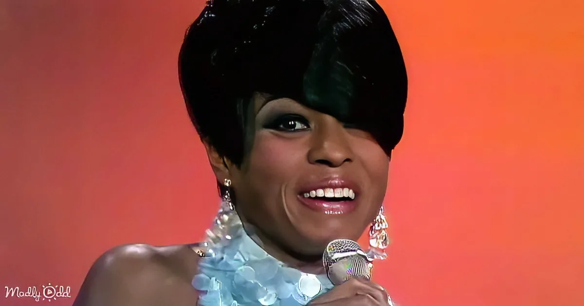 A person with short black hair, wearing a blue dress and earrings, holding a microphone against an orange background.