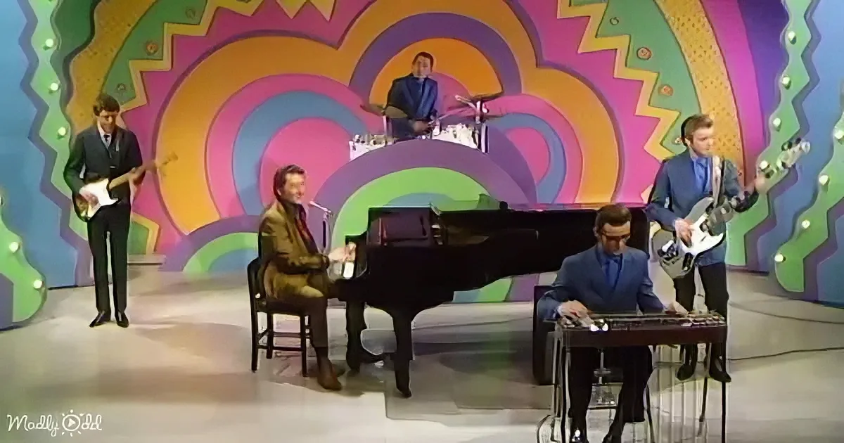 A band performs on stage with a colorful, psychedelic backdrop, featuring a pianist, keyboardist, guitarist, bassist, and...