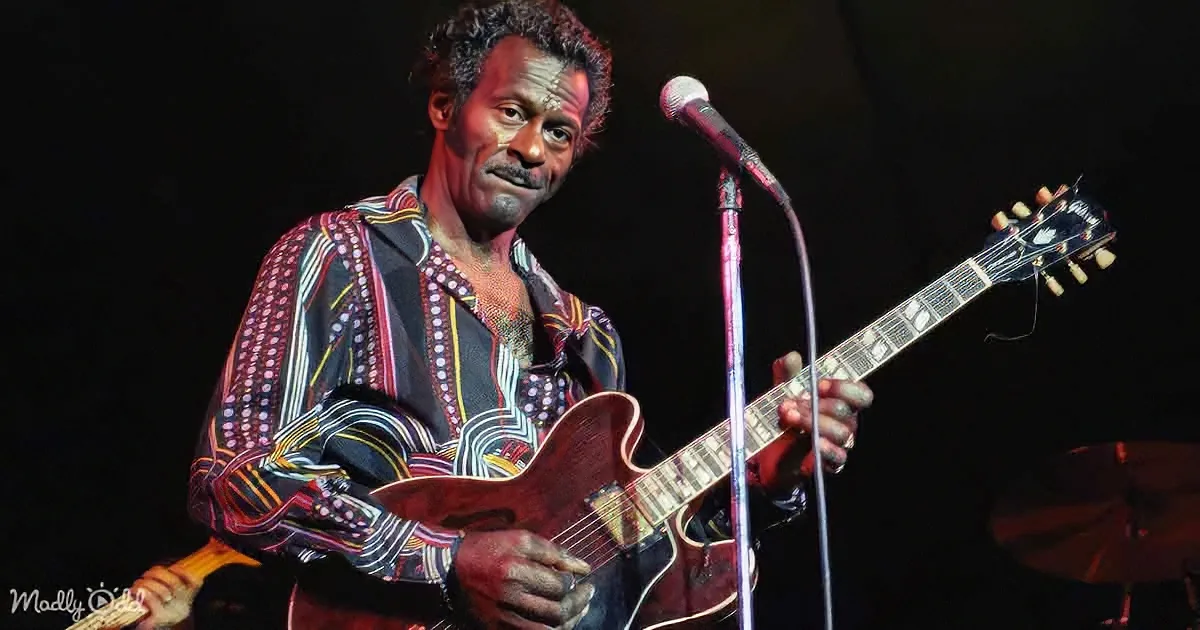 A man plays a guitar on stage, wearing a patterned shirt, with a microphone in front of him.