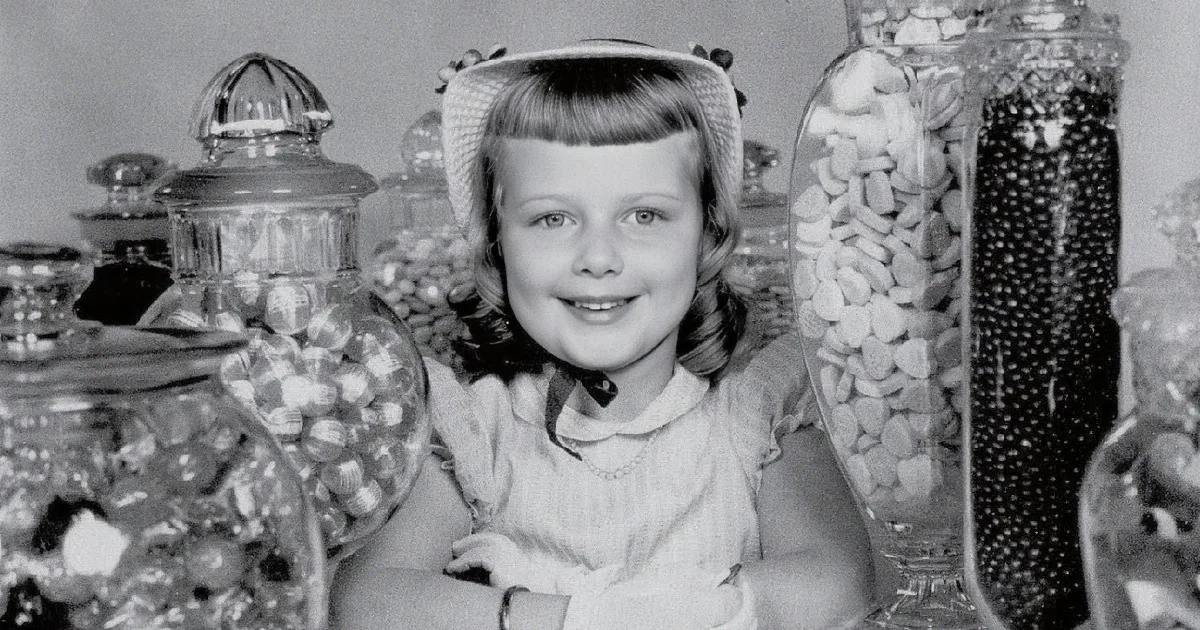 A man smiles as he hands a boy candy, with shelves of products and a sign for "Quality Candy" in the background.