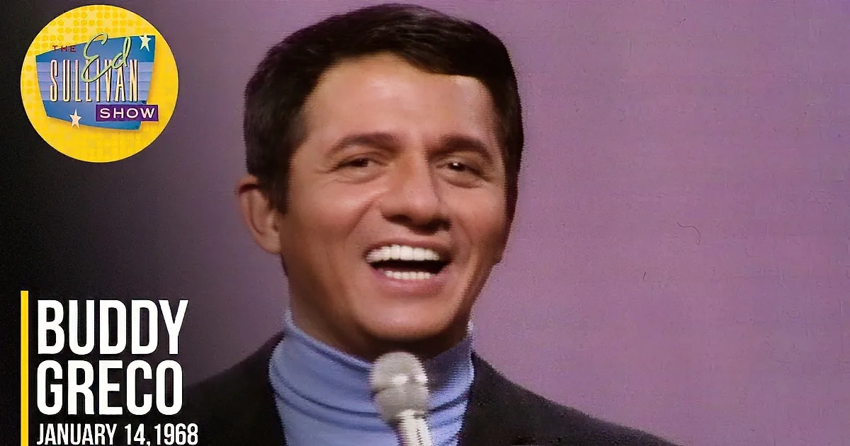 A man in a suit and blue turtleneck smiles while speaking into a microphone.