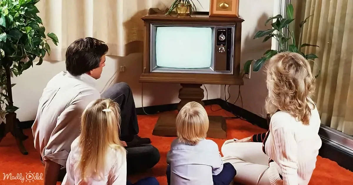 A family of four sits on the floor watching an old-fashioned television in a cozy living room.