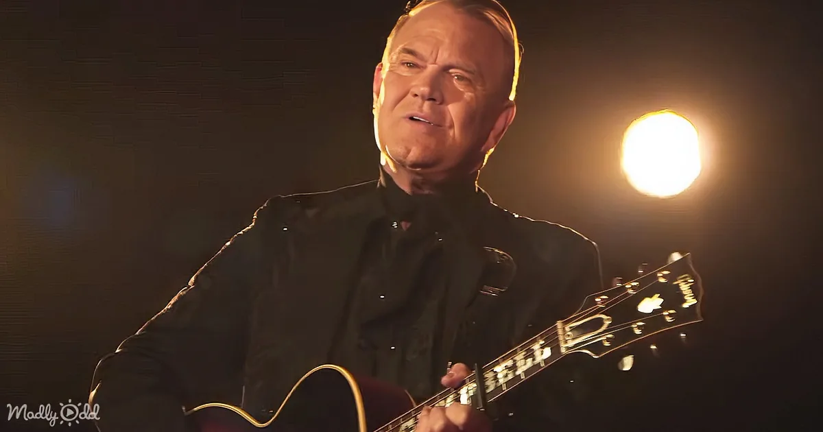 A man in a black suit plays an electric guitar under a spotlight.