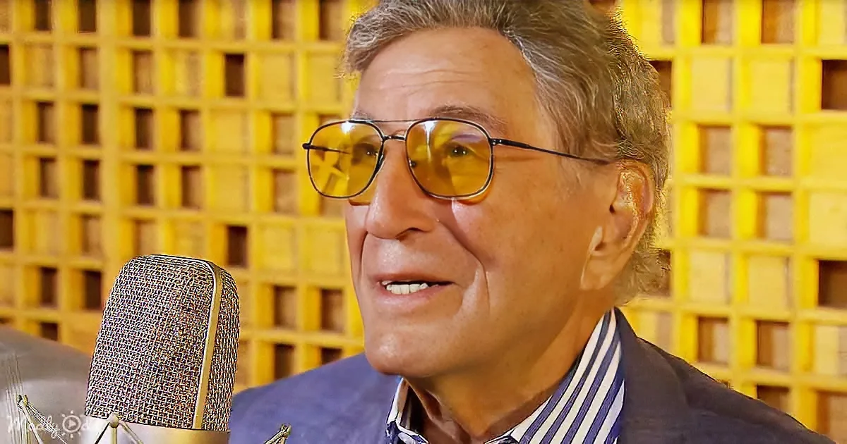 A man wearing aviator sunglasses and a striped shirt speaks into a microphone in front of a yellow acoustic foam background.