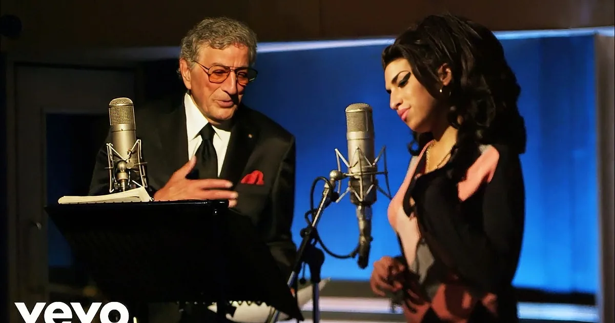 A man in a suit and a woman in a black dress are singing into microphones in a recording studio.