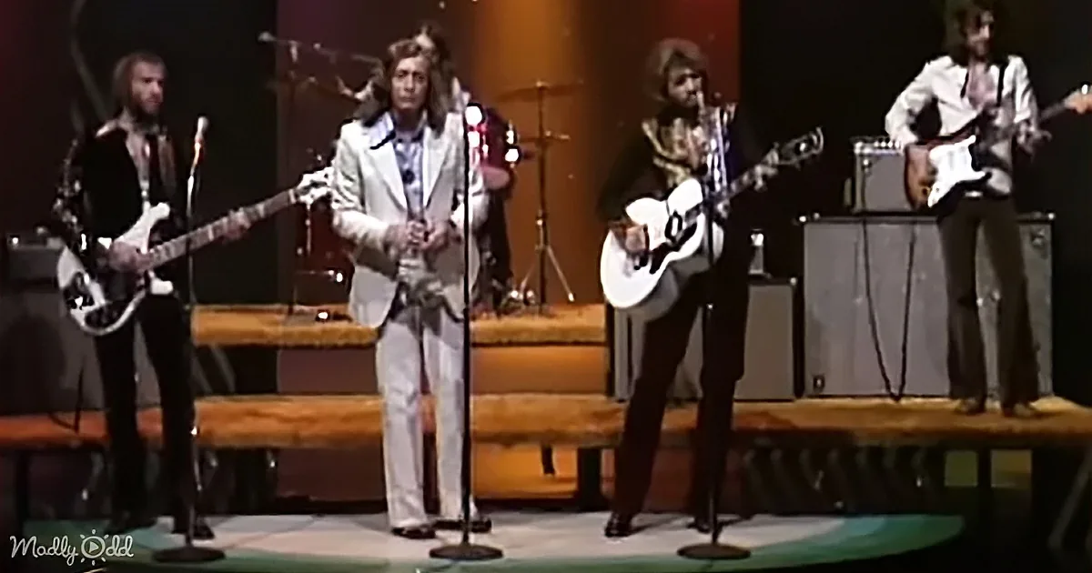 A band performs on stage with microphones, guitars, and a drum set, dressed in 1970s attire.