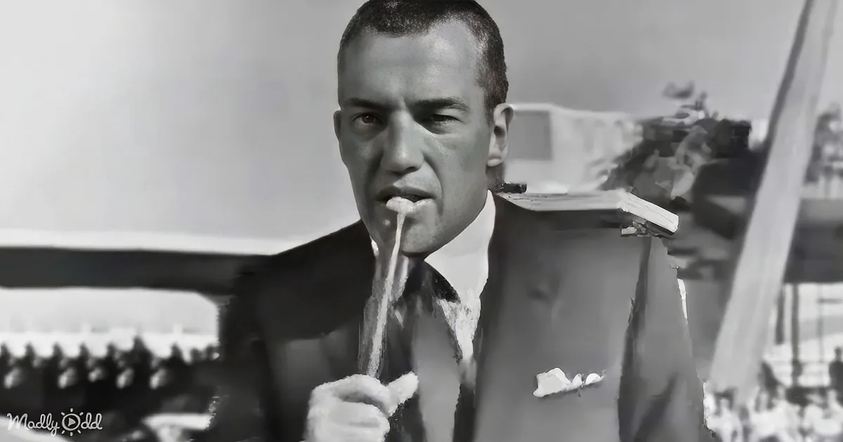 A man in a suit and tie is holding a microphone, with a blurred background of people and structures.