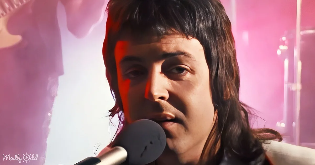 A person with long hair sings into a microphone against a pink-lit background.