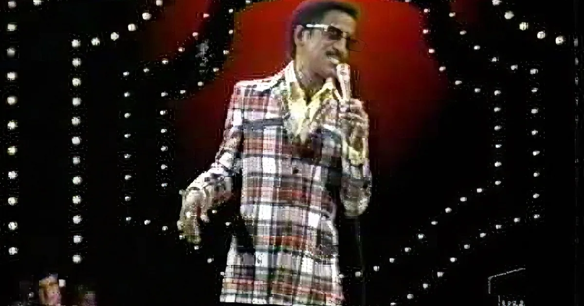 A man in a plaid shirt and sunglasses holds a microphone, singing against a red background with lights.