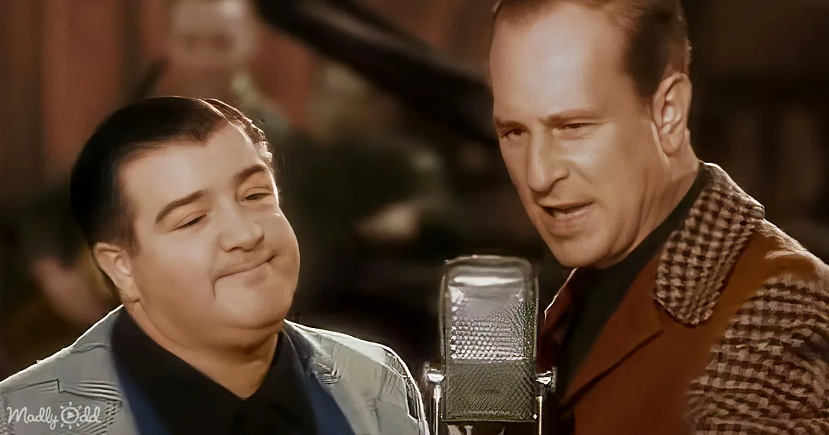 A man in a brown vest sings into a microphone, with another man in a blue jacket smiling beside him.