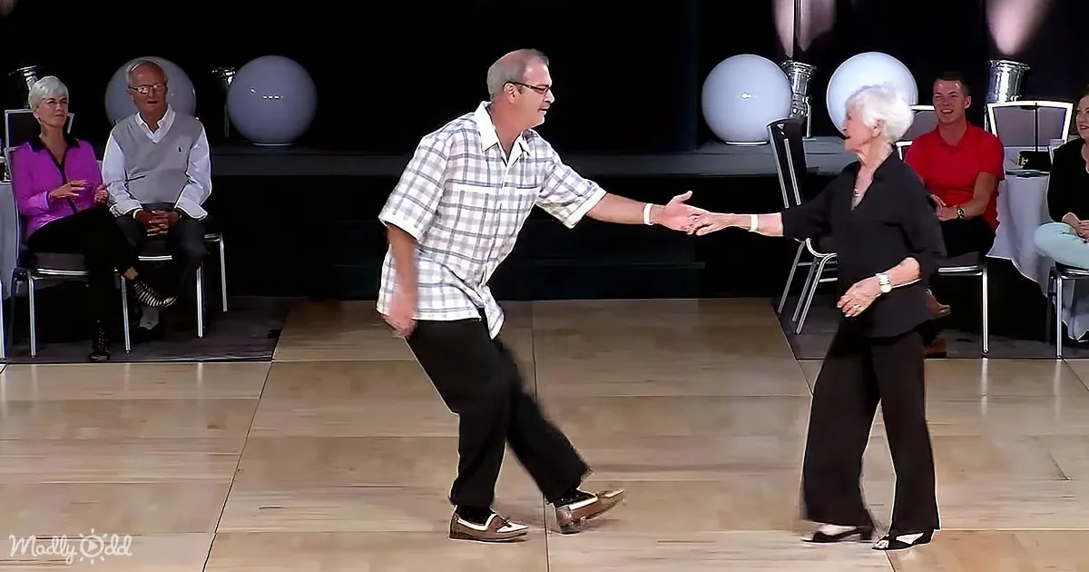 An older couple dances on a stage, holding hands, with spectators watching in the background.