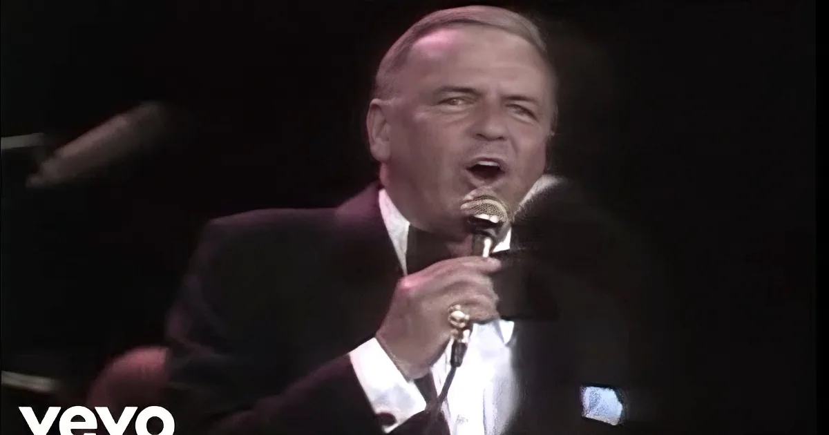 A man in a tuxedo sings into a microphone, smiling, with another person in the background.