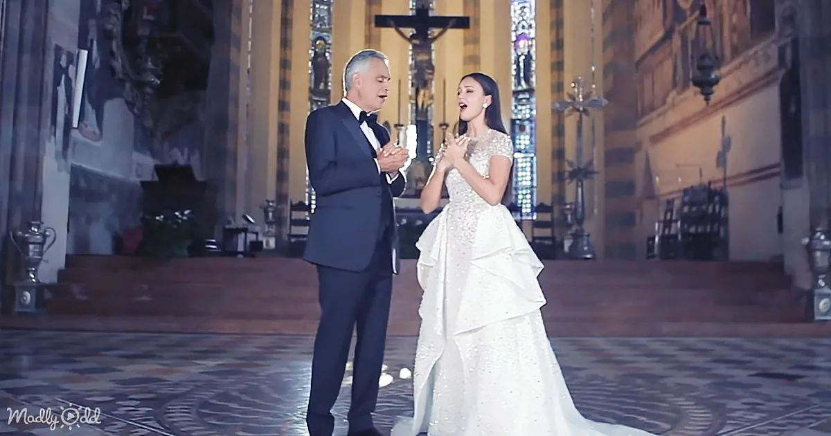 A man in a tuxedo and a woman in a white gown are singing on a stage with a church backdrop.
