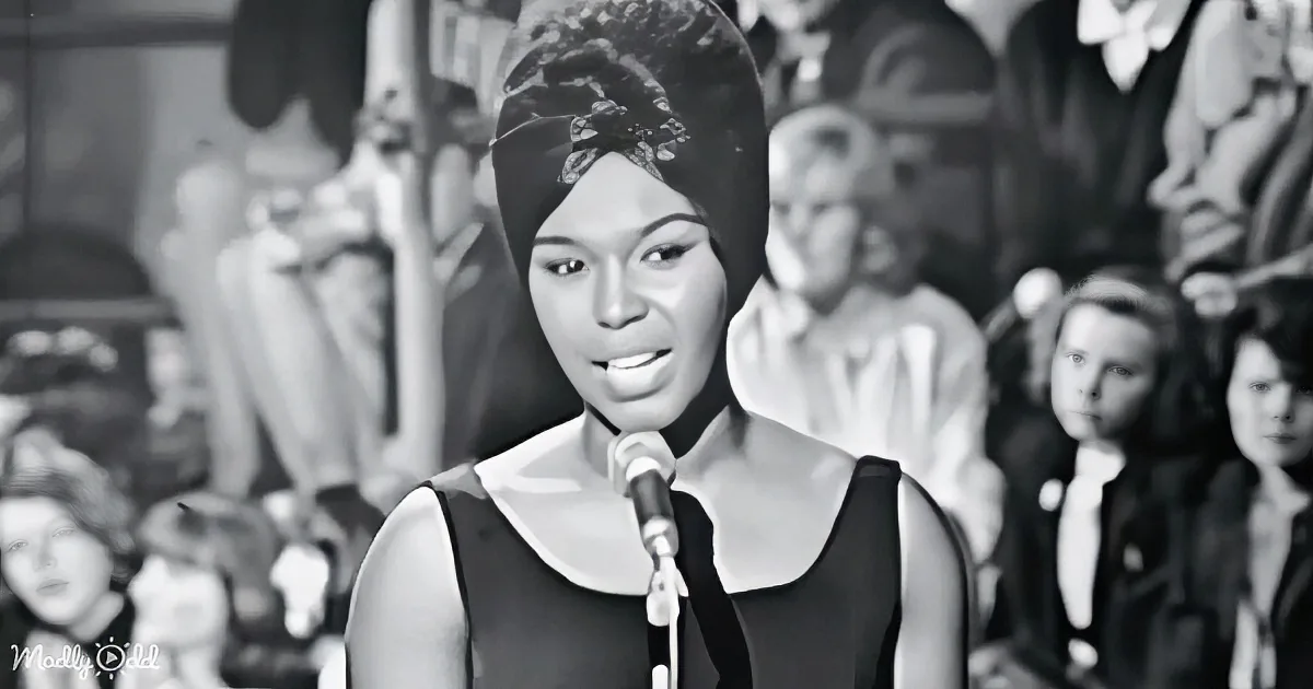 A woman in a black dress sings into a microphone, surrounded by a crowd in a vintage setting.