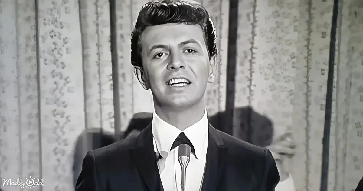 A man in a suit and tie sings into a microphone, smiling, with a patterned curtain in the background.