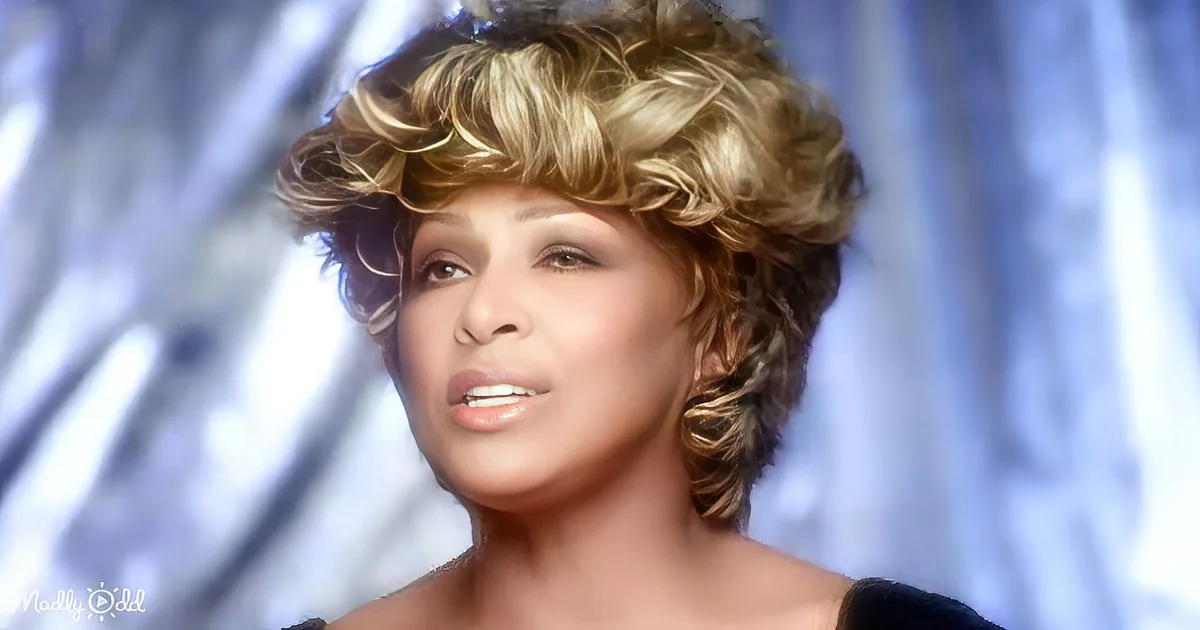 A woman with short, curly blonde hair and a light makeup look is shown against a blurred background.