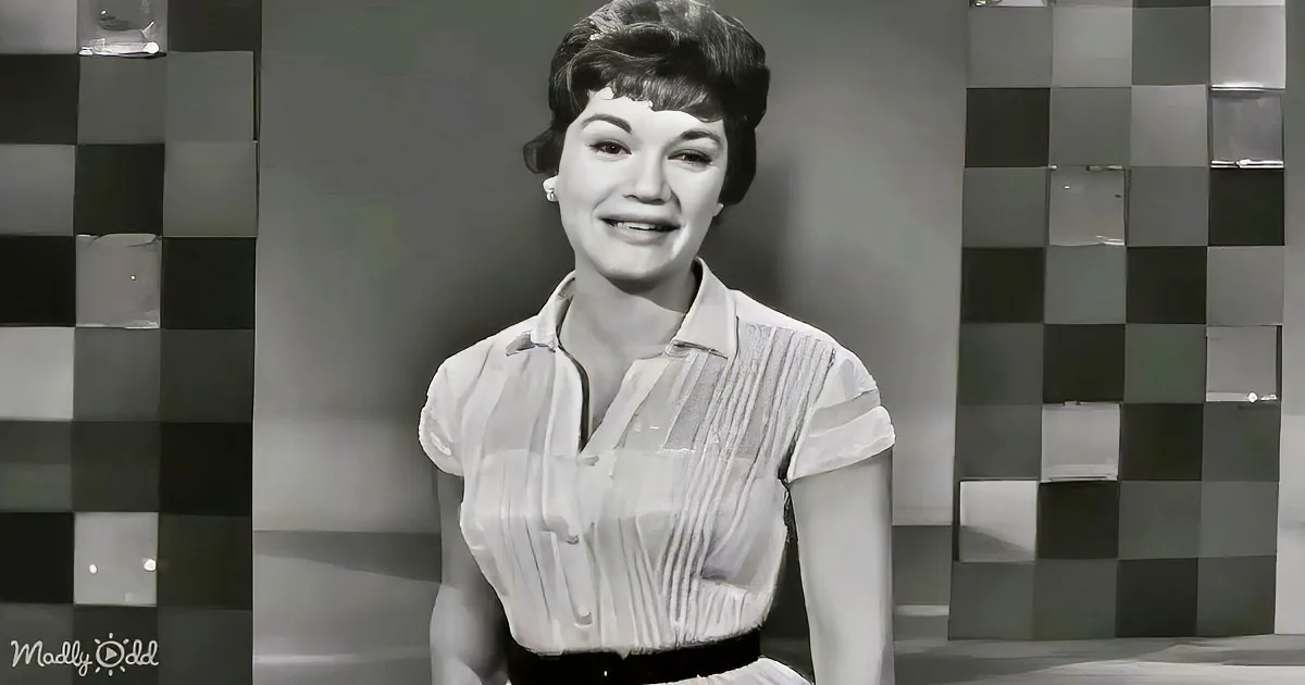 A woman in a white blouse smiles, standing against a checkered background.