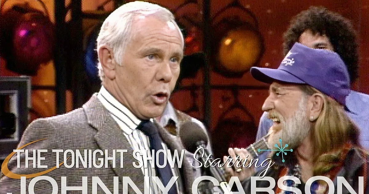 A man in a suit and striped shirt stands next to another man with a beard and a purple cap, both in front of a microphone...