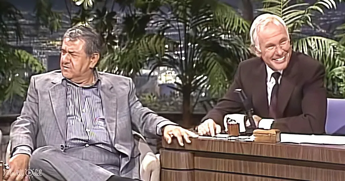Two men in suits, one seated at a desk smiling, the other in a chair, both in a talk show setting.