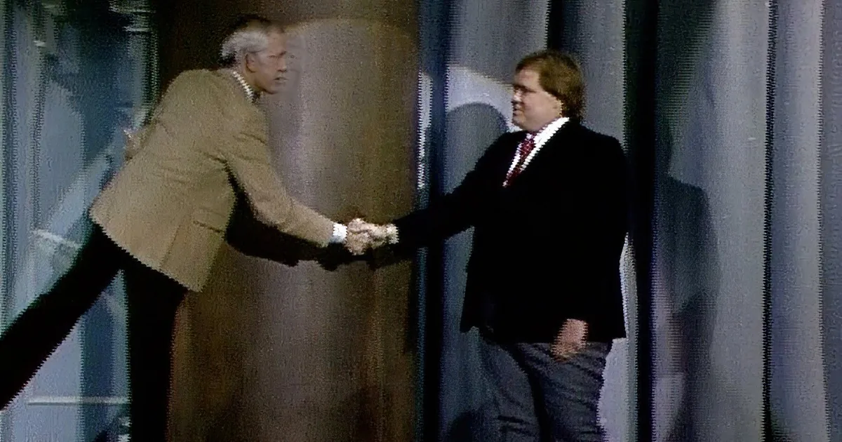 A man in a dark suit and red tie smiles widely, standing against a dark curtain backdrop.