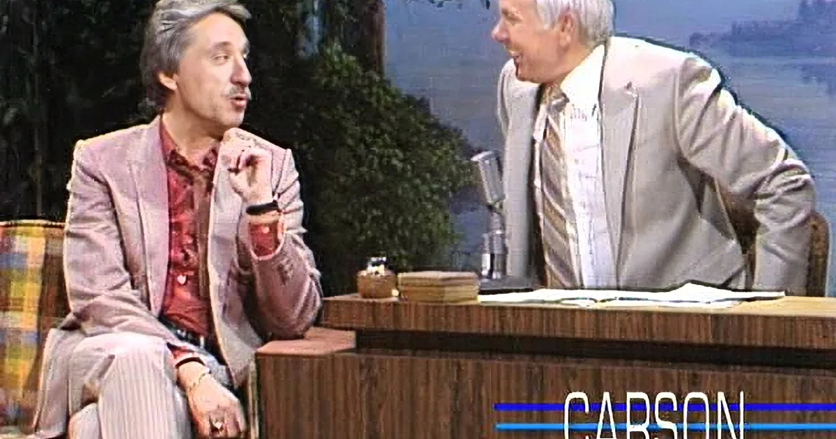 Two men in suits sit at a desk, laughing, with a scenic backdrop.