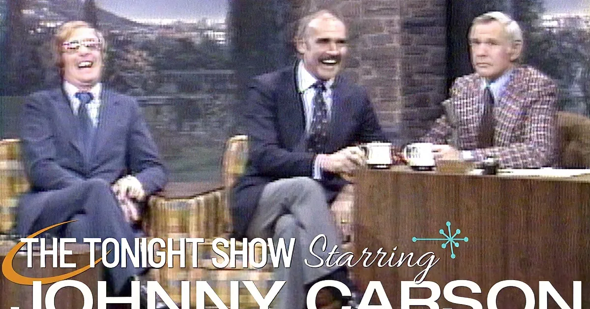 A man in a suit and glasses sits on a talk show set, with another man in a plaid jacket beside him.