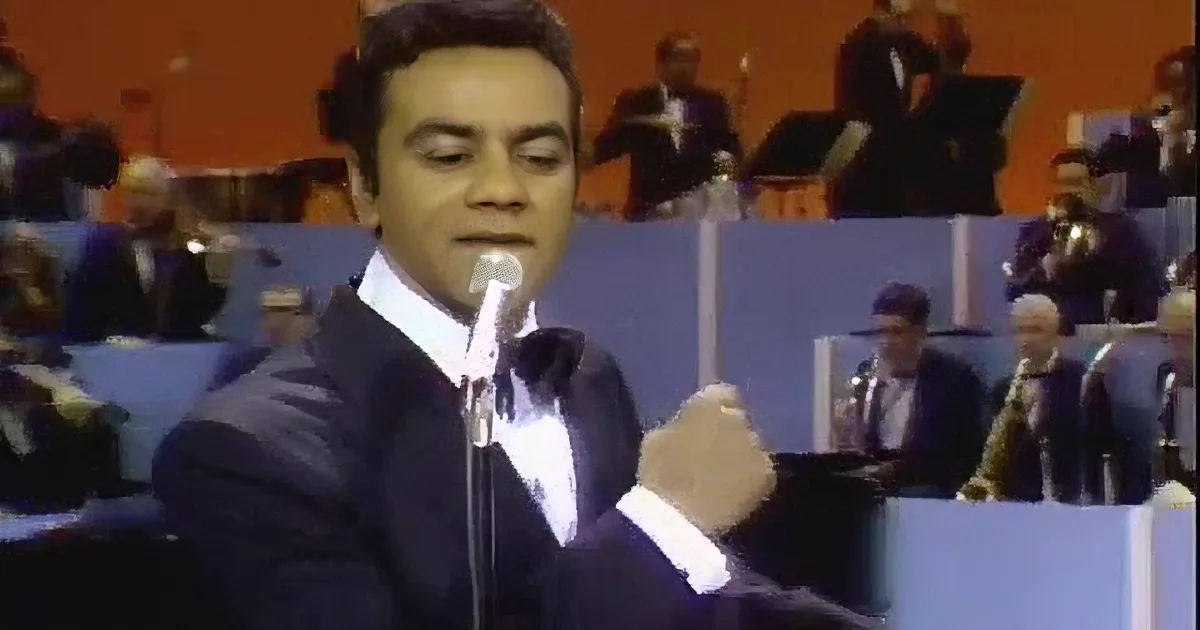 A man in a suit sings into a microphone on stage with an audience in the background.