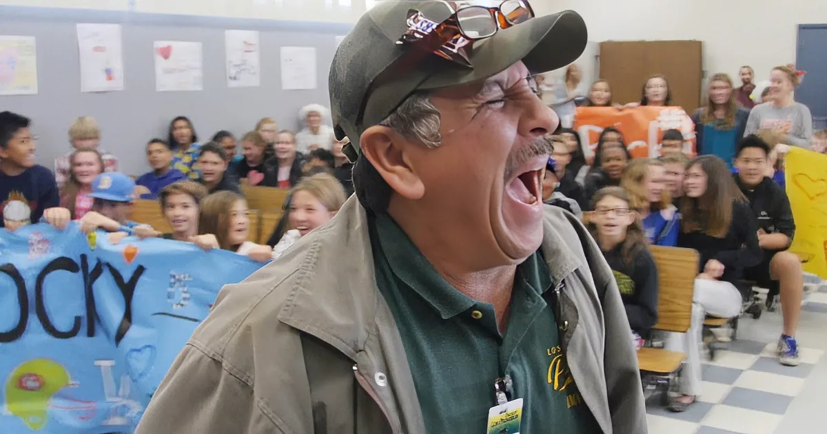 A man in a gray jacket stands with a group of people, some holding signs, in a room with a bucket of Gatorade and...