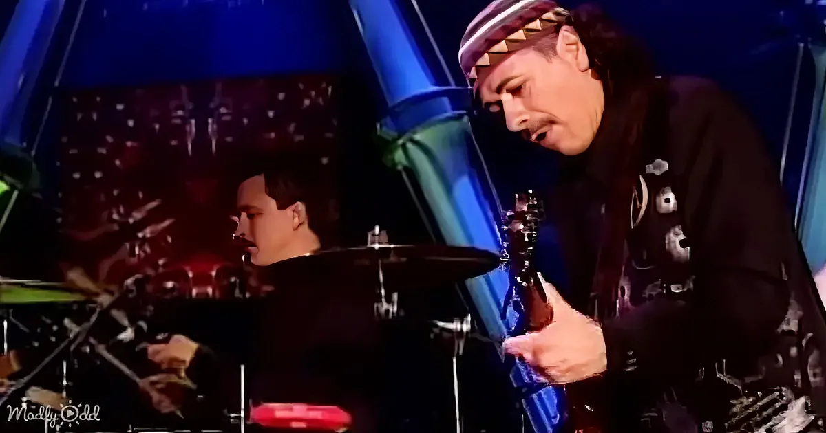 A guitarist in a black outfit with a spiked hat is playing an electric guitar on stage.