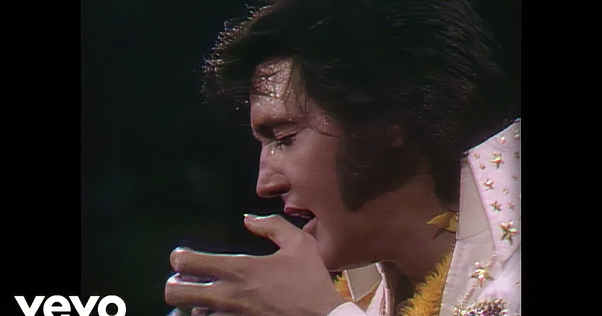 Elvis Presley in a white suit with yellow trim, singing into a microphone.