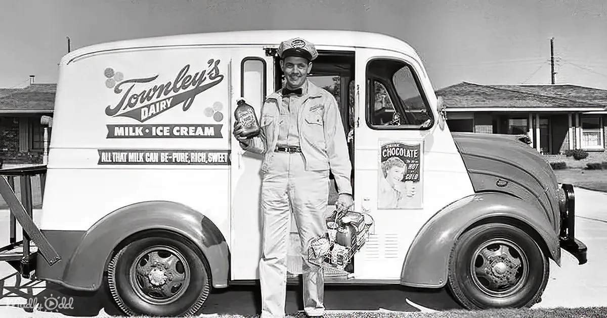 A milkman in uniform stands beside a vintage milk delivery truck, holding a milk bottle.