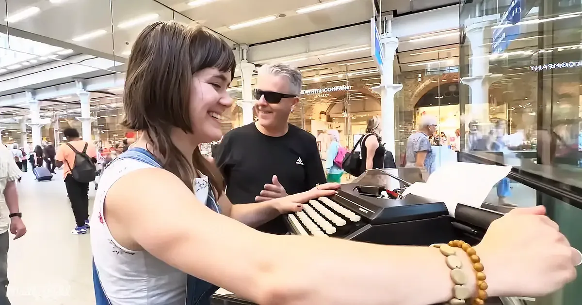 A woman smiles as she types on a vintage typewriter in a bustling shopping mall.