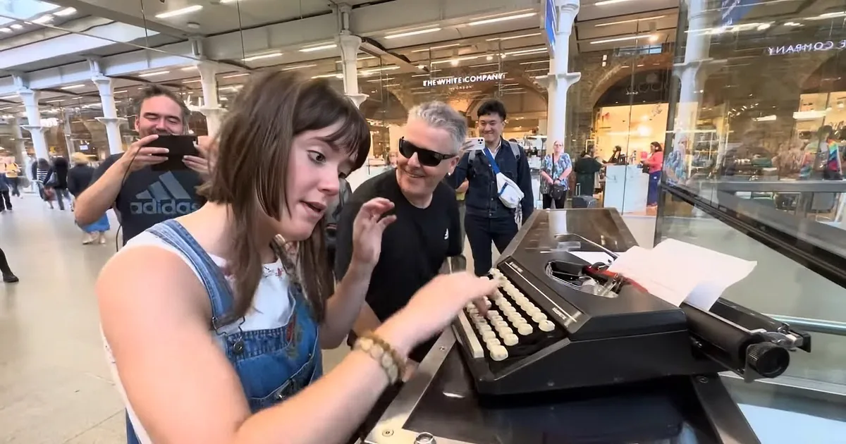 A woman smiles while typing on a vintage typewriter in a bustling shopping mall.