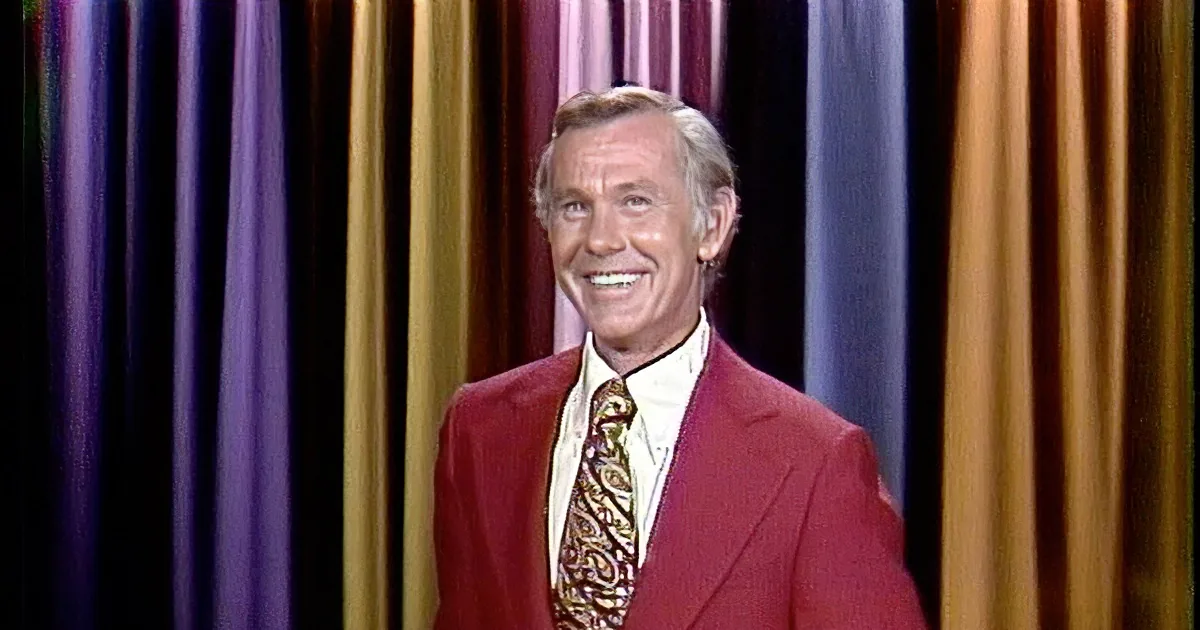 A man in a red suit and paisley tie smiles and gestures with his right hand.