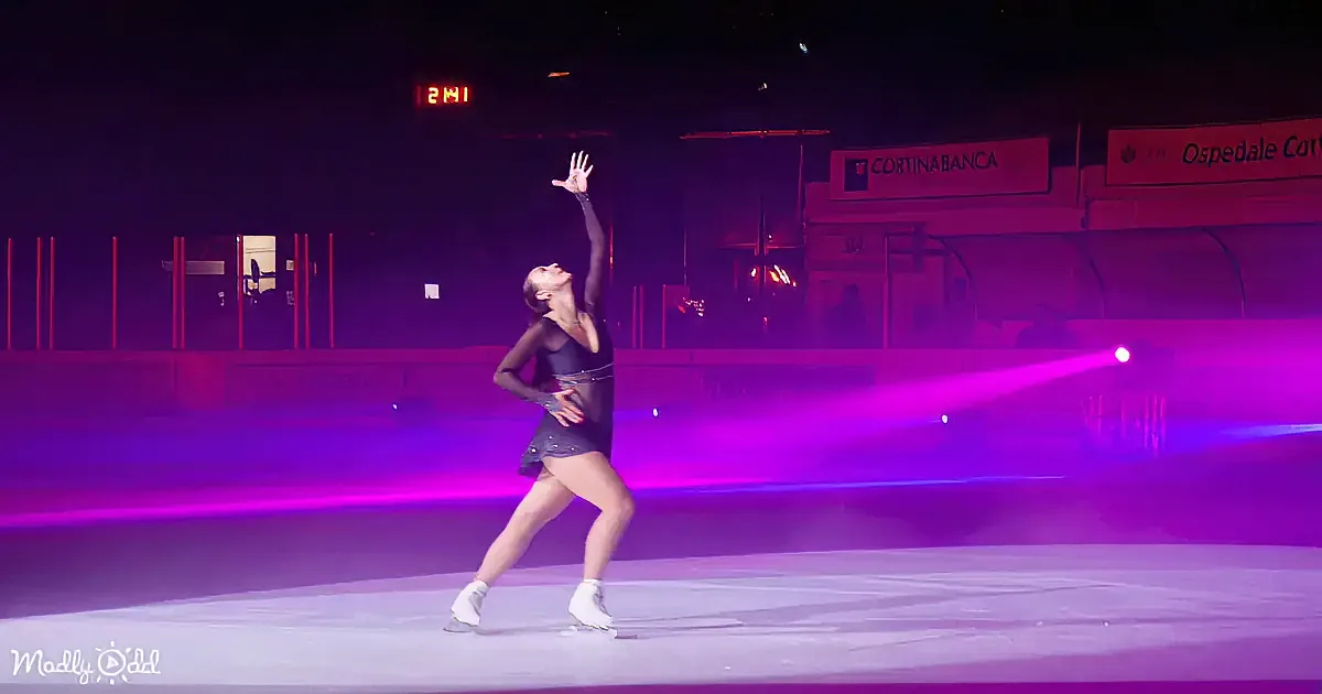 A figure skater in a black outfit performs a pose on ice with one arm raised, surrounded by purple lighting.