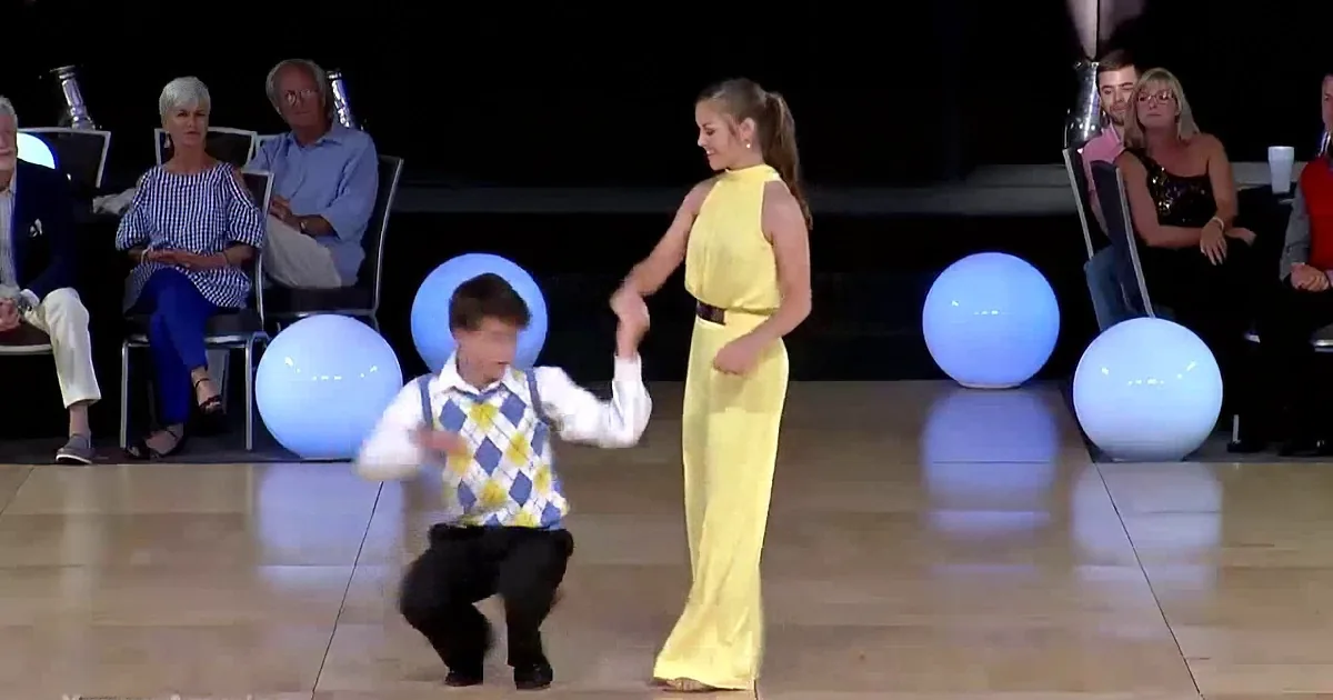 A couple in formal attire dances gracefully on a stage, surrounded by spectators and glowing orbs.