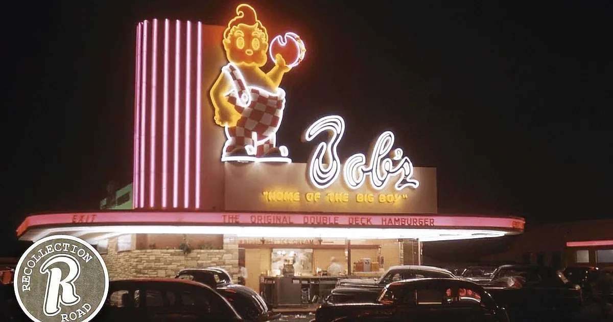 A brightly lit Bob's Big Boy sign with a cartoon character and neon lights, surrounded by cars in a parking lot at night.