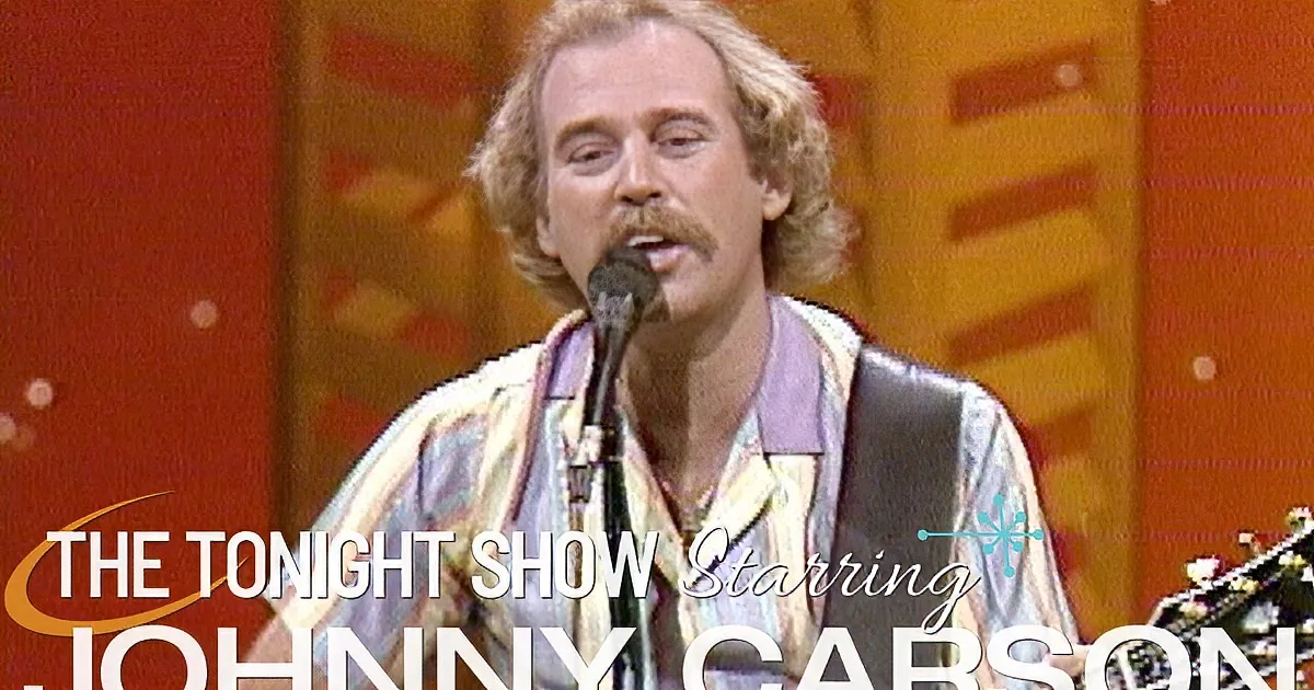 A man with a mustache and long hair sings into a microphone, wearing a colorful shirt and a guitar strap.