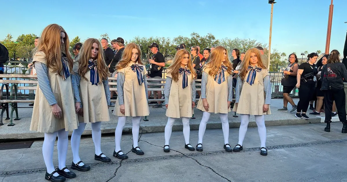 A group of performers in beige dresses and white tights dance in front of a crowd at a Halloween Horror Nights event.