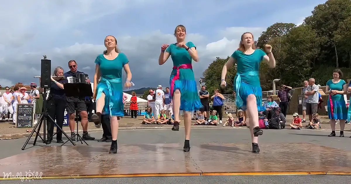 Three women in teal dresses dance on a stage while a band plays in the background.