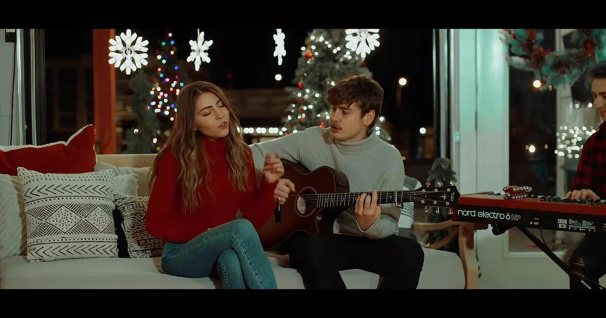 A man plays guitar while a woman sings in front of a Christmas tree.