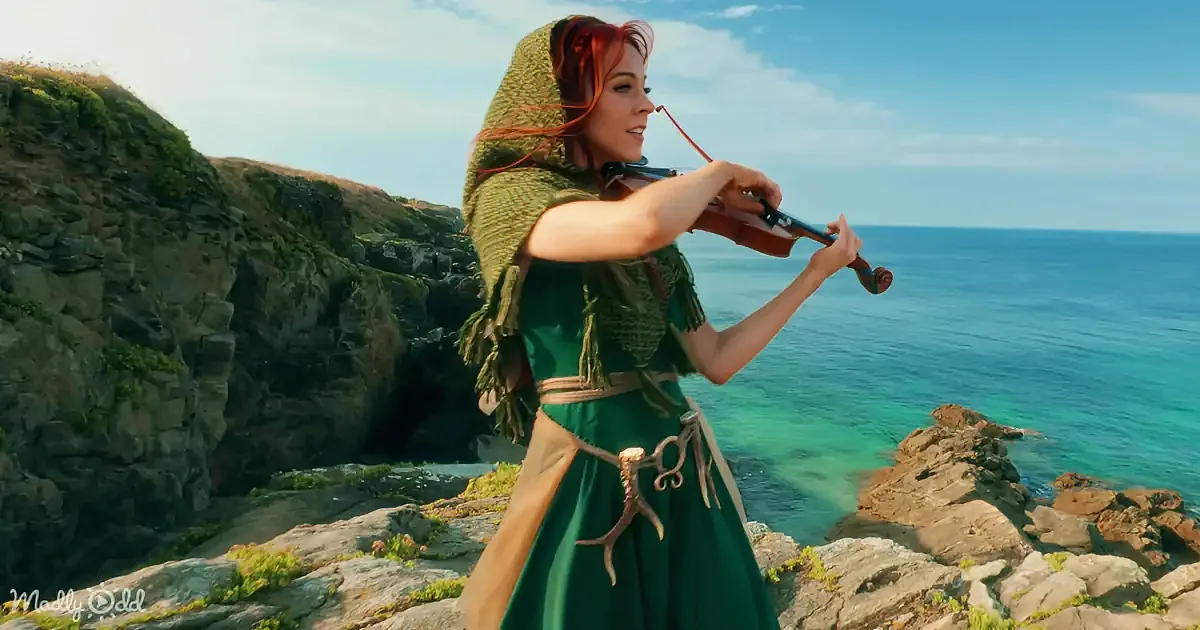 A woman in a green dress plays a violin on a rocky cliff overlooking the ocean.