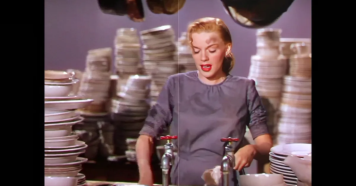 A woman with red lipstick and a polka-dot blouse leans on her hand, surrounded by stacks of plates and a large pot.