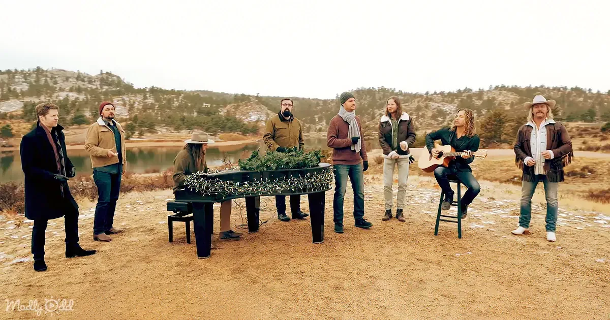 A group of seven people, including a guitarist and a pianist, are gathered outdoors near a lake, playing music.