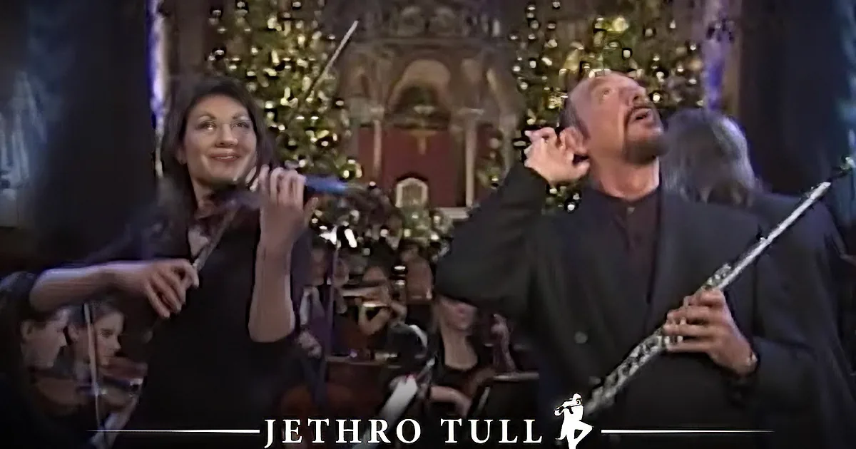 A man in a dark suit plays a flute in front of a decorated Christmas tree.